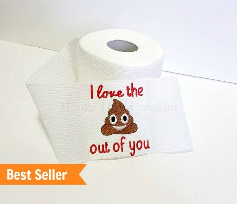 Op de afbeelding: Een witte toiletrol met rode borduurwerk die "I love the out of you" zegt met een bruine poep-emoji met een smiley.