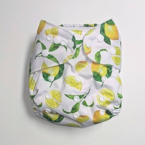 May include: A white cloth diaper with a lemon print. The diaper has white snaps and a white elastic waistband.