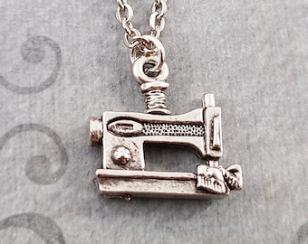 May include: Silver-colored sewing machine pendant necklace. The pendant is a detailed miniature of a vintage sewing machine, suspended from a delicate chain. The necklace is displayed against a gray background with a subtle pattern.