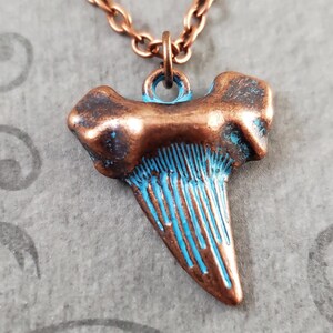 May include: Copper shark tooth necklace with a blue patina finish. The tooth is detailed with lines and has a rustic look.