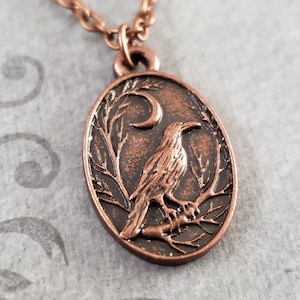 May include: A copper-colored oval pendant necklace featuring a detailed raven perched on a branch, with a crescent moon in the background. The pendant is framed by a decorative border and hangs from a matching chain. The overall design has a vintage or gothic aesthetic.
