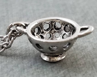 Op de afbeelding: Een zilveren zeefhangerketting. De zeef is een klein, gedetailleerd replica van een keukenzeef met een handvat. De ketting is gemaakt van een zilveren ketting.