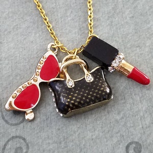 May include: Gold-tone necklace with charms including red sunglasses, a black handbag, and a red lipstick. The sunglasses have gold frames and red lenses. The handbag is black with gold accents and the lipstick has a red tube.