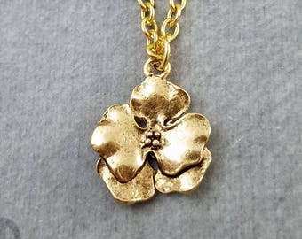 May include: A gold-colored necklace featuring a three-petal flower pendant. The pendant has a textured surface and a small cluster of beads at its center. The chain is also gold-colored and has a link design. The background is a solid gray color. The text "CharmsOnThings" is visible in the lower left corner.