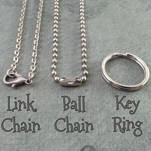 Puede incluir: Tres tipos diferentes de cadenas: una cadena de eslabones plateada, una cadena de bolas plateada y un llavero plateado. Las cadenas est&aacute;n dispuestas sobre una superficie gris texturizada.