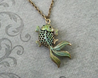May include: A gold-tone necklace with a green and gold fish pendant. The fish has a detailed, scaled design and a long, flowing tail.
