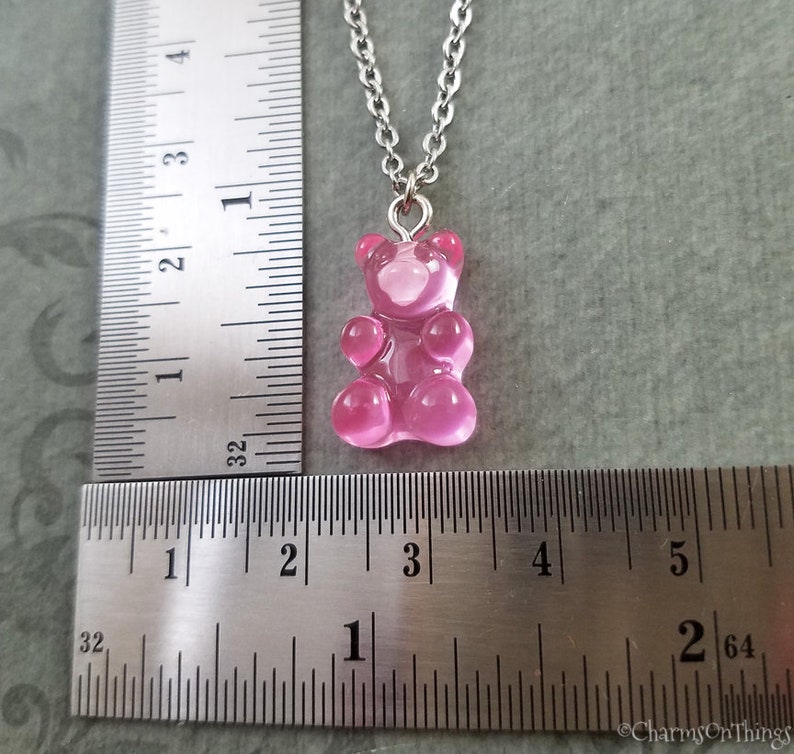 Puede incluir: Un collar con un dije de osito de gomita rosa y una cadena de plata. El dije mide aproximadamente 2,5 cm de alto y est&aacute; colgado de una regla.