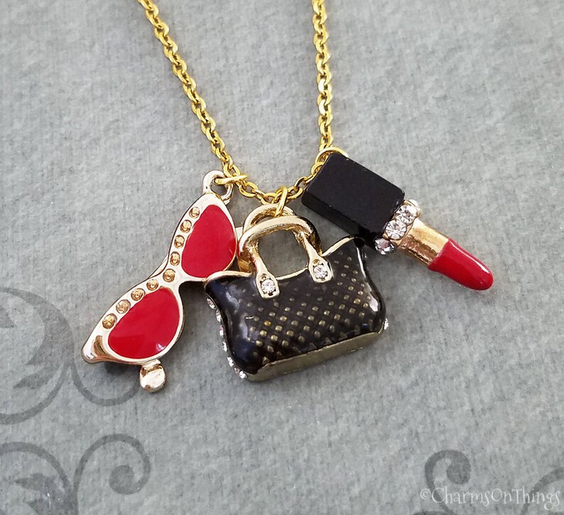 May include: Gold-tone necklace with charms including red sunglasses, a black handbag, and a red lipstick. The sunglasses have gold frames and red lenses. The handbag is black with gold accents and the lipstick has a red tube.