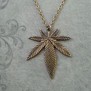 May include: A gold-toned necklace featuring a large, detailed marijuana leaf pendant. The pendant is crafted from metal and has a realistic design.