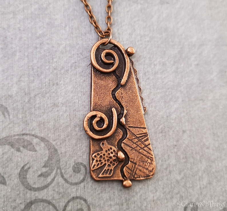 May include: A copper pendant necklace with a textured, abstract design featuring swirls, lines, and a bird. The pendant is attached to a copper chain.