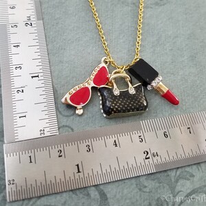 May include: Gold-tone necklace with charms including red sunglasses, a black handbag, and a red lipstick. The sunglasses have rhinestone accents. The handbag and lipstick also have rhinestone accents. The necklace is approximately 18 inches long.