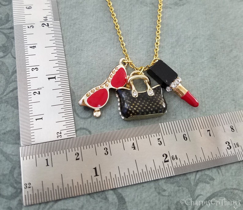 May include: Gold-tone necklace with charms including red sunglasses, a black handbag, and a red lipstick. The sunglasses have rhinestone accents. The handbag and lipstick also have rhinestone accents. The necklace is approximately 18 inches long.