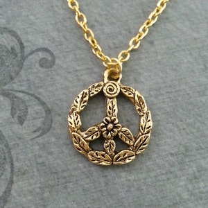 May include: A gold-colored necklace featuring a peace symbol pendant. The pendant is a detailed design with a floral element at the center and a wreath of leaves around the peace symbol. The chain is also gold-colored.