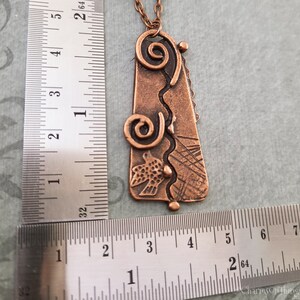 May include: A copper pendant necklace with a textured, abstract design featuring a bird and swirls. The pendant is approximately 2 inches long and is shown next to a ruler for size reference.