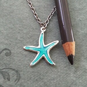May include: A silver necklace featuring a turquoise starfish pendant. The starfish has a silver border. The chain is silver. A dark grey pencil is placed beside the necklace for scale.