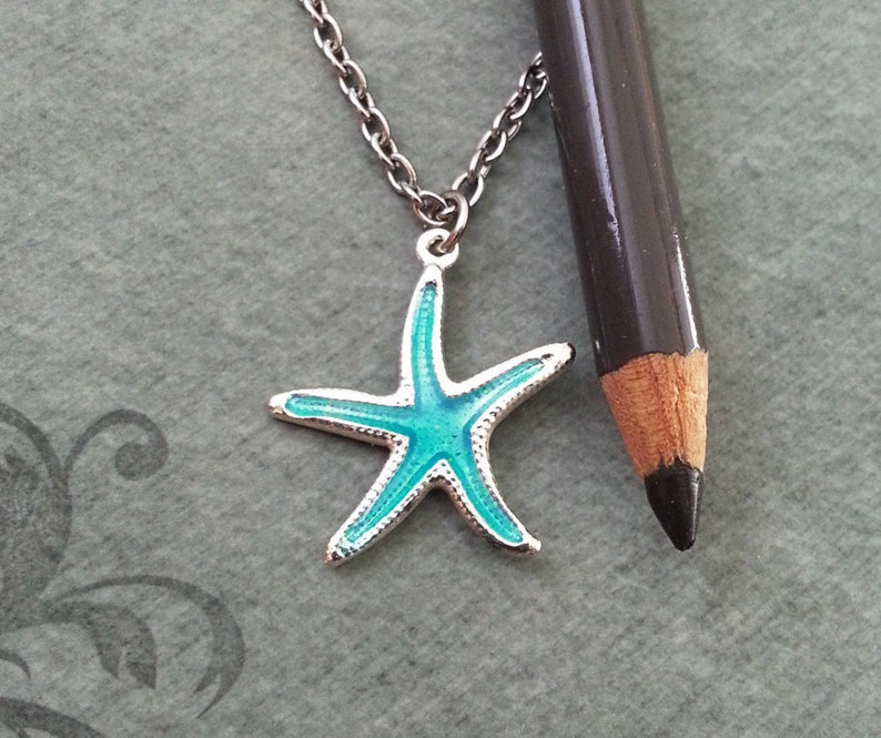 May include: A silver necklace featuring a turquoise starfish pendant. The starfish has a silver border. The chain is silver. A dark grey pencil is placed beside the necklace for scale.