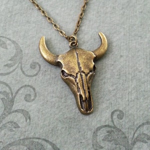 May include: A bronze-colored pendant necklace featuring a detailed bull skull design. The pendant is attached to a chain.