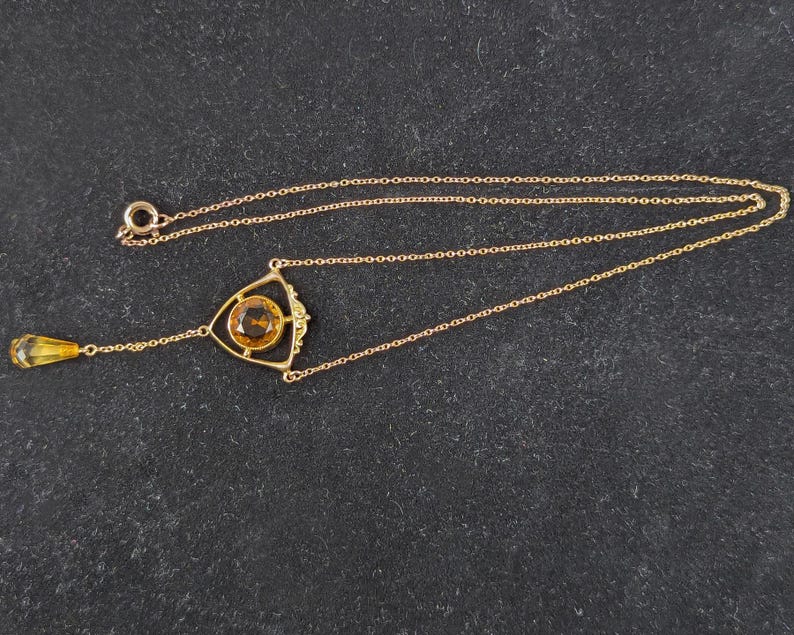 May include: A gold necklace with a teardrop-shaped pendant and a triangular pendant with a round, orange gemstone.