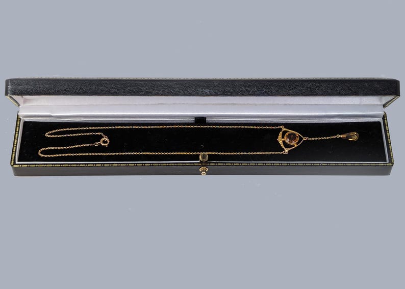 May include: A gold necklace with a pendant featuring a round, dark brown stone set in a delicate, geometric design. The necklace is displayed in a black velvet jewelry box.