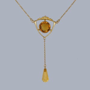 May include: A gold necklace with a teardrop-shaped pendant and a triangular centerpiece featuring a round, amber-colored gemstone.