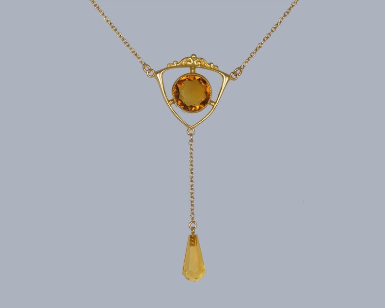 May include: A gold necklace with a teardrop-shaped pendant and a triangular centerpiece featuring a round, amber-colored gemstone.