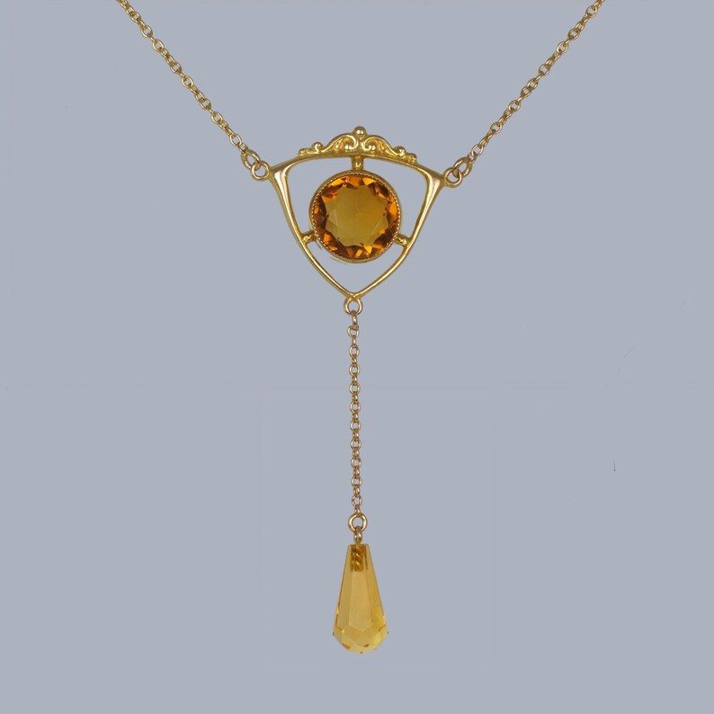 May include: A gold necklace with a teardrop-shaped pendant and a triangular centerpiece featuring a round, amber-colored gemstone.