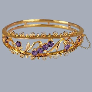 以下が含まれることがあります： 金色のヒンジ付きバングルブレスレット。紫色の宝石と、花と渦巻きの装飾が施されています。小さなチェーンで留められています。ジュエリー。