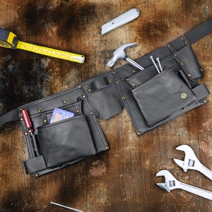 Pode incluir: Um cinto de ferramentas de couro preto com vários bolsos e um martelo, uma chave de fenda e pregos. O cinto é fechado com uma fivela e tem rebites de metal.