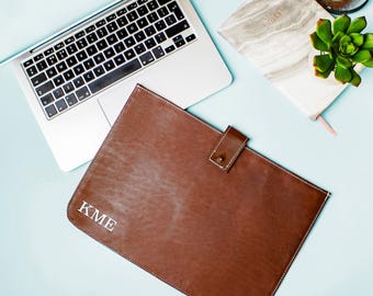 Puede incluir: Una funda para portátil de cuero marrón con un monograma blanco "KME" y un cierre de botón dorado. La funda está sobre una superficie azul claro.