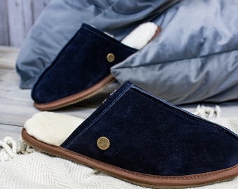 May include: A pair of navy blue suede slippers with a brown leather trim and a sheepskin lining. The slippers have a gold metal logo on the top.
