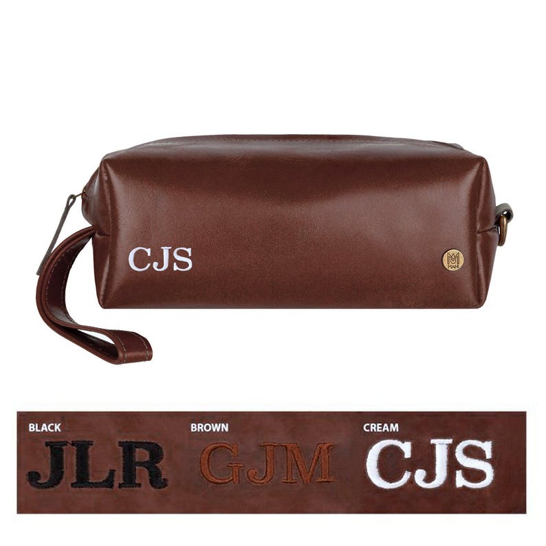 Puede incluir: Bolsa de aseo de viaje de cuero marr&oacute;n con un monograma blanco "CJS" y un peque&ntilde;o logotipo dorado. La bolsa tiene una cremallera y un asa.