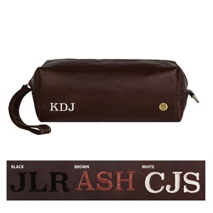 Puede incluir: Una bolsa de viaje de cuero marr&oacute;n con un monograma blanco "KDJ" y una etiqueta de metal dorado. La bolsa tiene una cremallera y un asa.