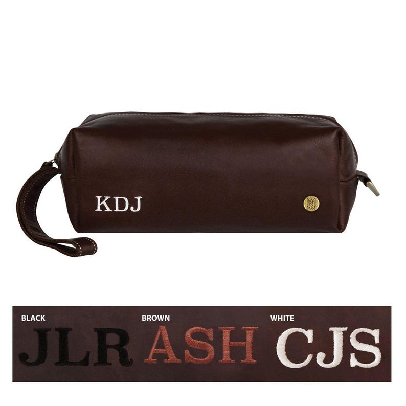 Puede incluir: Una bolsa de viaje de cuero marr&oacute;n con un monograma blanco "KDJ" y una etiqueta de metal dorado. La bolsa tiene una cremallera y un asa.