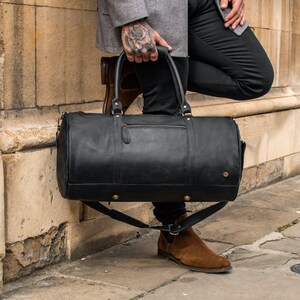 May include: A black leather duffel bag with gold hardware and a long strap. The bag is being held by a person wearing black pants and brown boots.