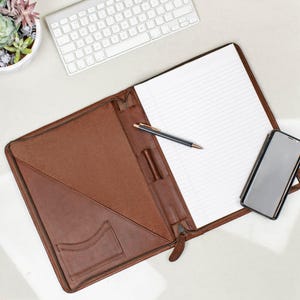 May include: A brown leather portfolio with a zipper closure, a pen holder, and a pocket for a phone. The portfolio is open and has a lined notepad inside.