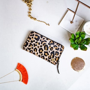 May include: A leopard print wallet with a gold zipper pull and a small gold circle on the front. The wallet is on a white surface with a gold necklace, a small succulent plant, and a compact mirror.