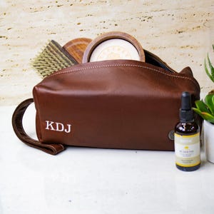 May include: A brown leather toiletry bag with a zipper closure and a white embroidered monogram "KDJ". The bag is open and contains a wooden bowl, a brush, and a bottle of beard oil.