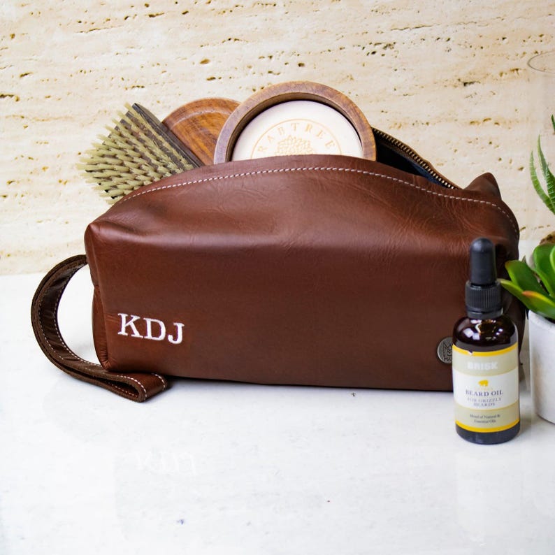 May include: A brown leather toiletry bag with a zipper closure and a white embroidered monogram "KDJ". The bag is open and contains a wooden bowl, a brush, and a bottle of beard oil.