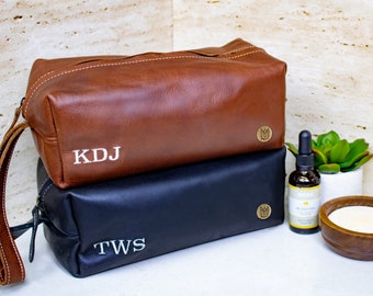 Puede incluir: Dos bolsas de aseo de cuero, una marrón y otra negra, apiladas una encima de la otra. La bolsa marrón tiene las iniciales "KDJ" bordadas, y la bolsa negra tiene las iniciales "TWS" bordadas. Ambas bolsas tienen una pequeña etiqueta de latón con un logotipo.