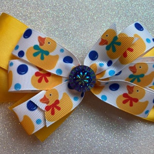 May include: A yellow hair bow with a white ribbon featuring a pattern of yellow rubber ducks and blue polka dots. The bow is centered with a dark blue jewel.