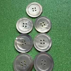 May include: Seven circular buttons with a pearlescent, off-white finish. Each button features a smooth, slightly concave center and a raised outer rim, with four small holes arranged in a square pattern in the middle for attachment to fabric.