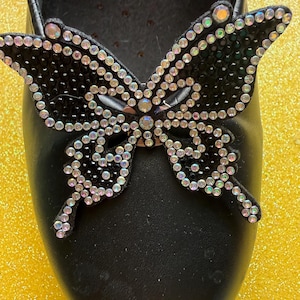 May include: Black shoe with a large butterfly embellishment. The butterfly is black and covered in iridescent rhinestones. The shoe is made of a smooth, shiny material.