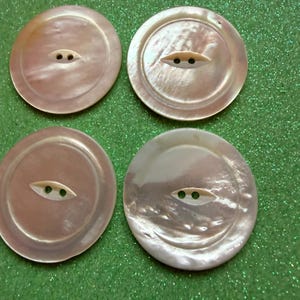 May include: Four round buttons made of iridescent mother-of-pearl material. Each button features a smooth, polished surface with a subtle pinkish hue and a central, recessed eye-shaped detail containing two small holes for sewing attachment.