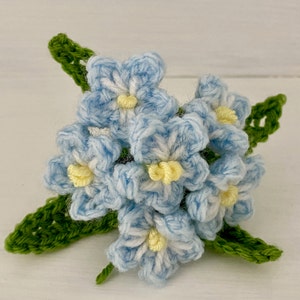 May include: A crocheted bouquet of blue forget-me-not flowers with yellow centers. The flowers are arranged in a cluster with green leaves.