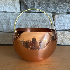 May include: A copper planter with a brass handle. The planter is round and has a reflective surface.