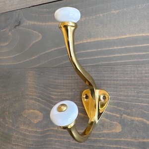 May include: A gold metal coat hook with two white ceramic knobs. The hook is curved and has a heart-shaped base.