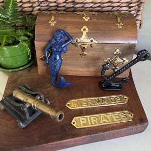 May include: A collection of nautical-themed decor items. Includes a wooden treasure chest with brass accents, a blue mermaid figurine, a black anchor, a brass cannon, and brass plaques that read "MERMAIDS" and "PIRATES".