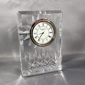 May include: A Waterford crystal clock with a clear glass base and a silver face with black Roman numeral markings. The clock face reads "Waterford Crystal".