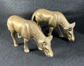 May include: Two brass horse figurines, both standing on a black surface. The horses are facing the same direction, with their heads lowered as if they are grazing.