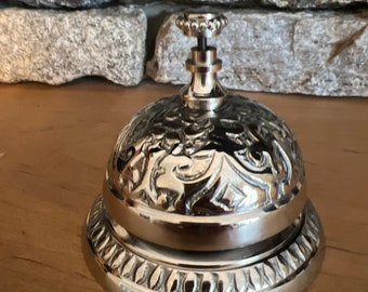 May include: A silver-toned bell with an ornate design. The bell has a round top and a base with a decorative pattern. The bell is made of metal and has a polished finish.